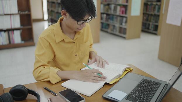 College student writing and reviewing notes with a laptop in a library setting